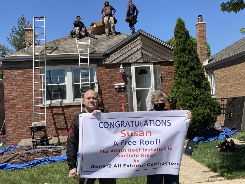 With an All Exterior Contractors crew busy on her roof above, Garfield Ridge homeowner Susan Donner (right) holds her congratulatory banner with AEC owner Geno Randazzo. --Photo by Tim Hadac